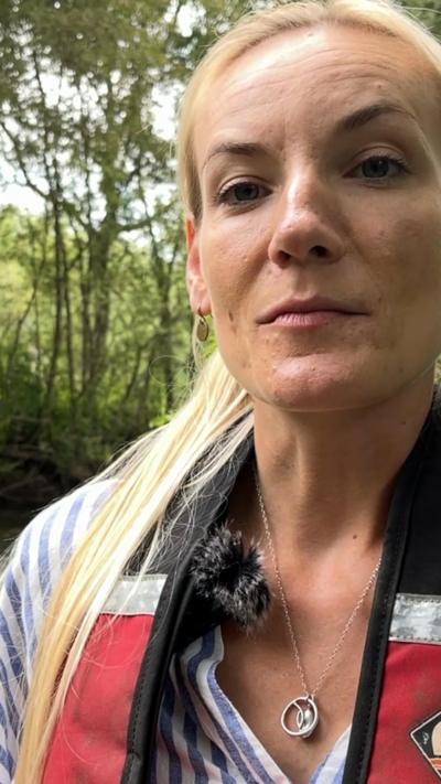 A woman wearing a life jacket is standing in a stream, she has blonde hair.