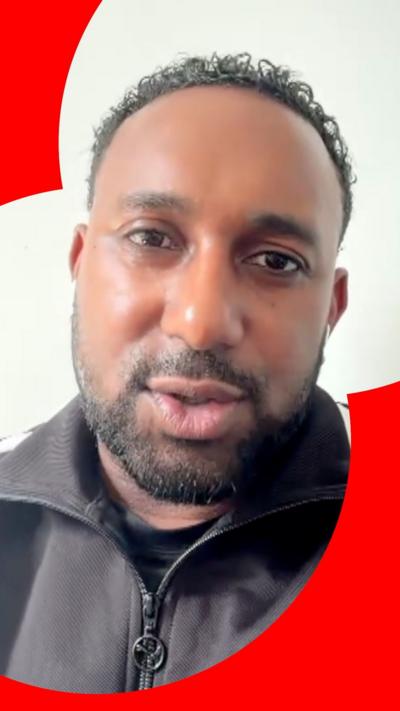 A man with short black hair, moustache and beard talks to the camera. He is wearing a zipped up sports jacket.