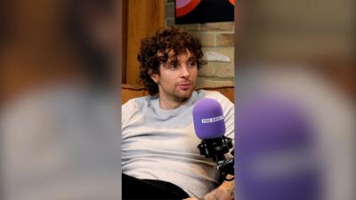 Tom Grennan sits on a couch wearing a grey T-shirt behind a purple microphone