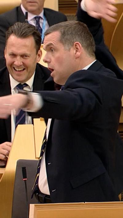 A man in a suit waves his arms around in the Scottish Parliament chamber.