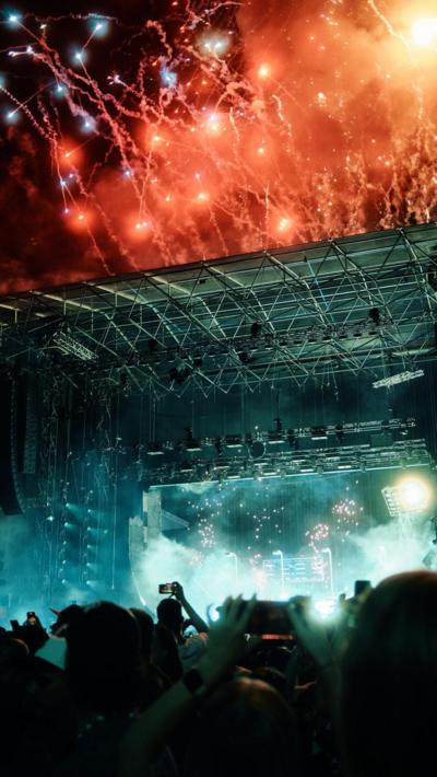 A crowd in front of a large stage with red fireworks going off above it. 