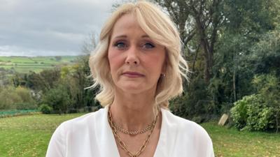 Deirdre Shakespeare wearing a white blouse and gold necklace. She has wavy blonde hair and is outside. There are trees and fields behind her.