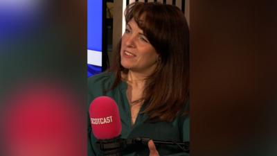 Catriona MacPhee in a green shirt speaking into a pink microphone with the word 'Scotcast' on it.