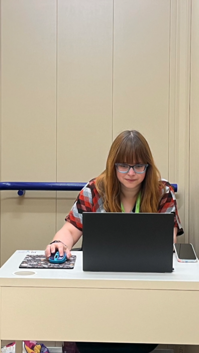 A woman with brown hair and a fringe sitting behind a laptop at a desk. She has set up her own personal office inside a lift.