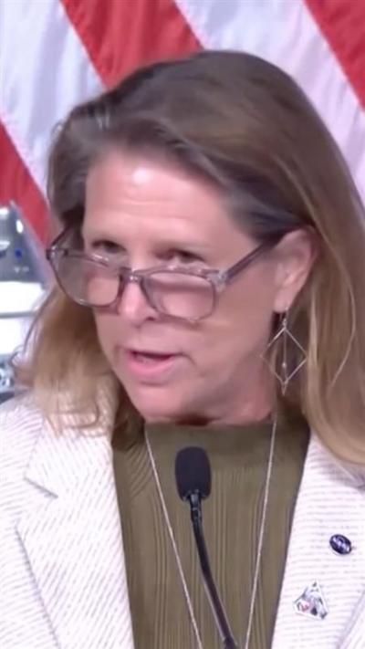 A woman in glasses speaks into a microphone as the red and white stripes of a US flag hang behind her.