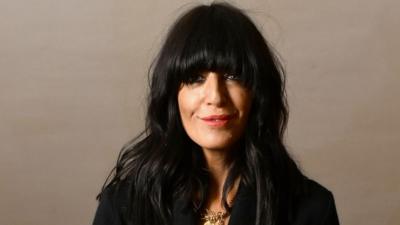Claudia Winkleman wearing black clothes and gold necklace looking at camera.