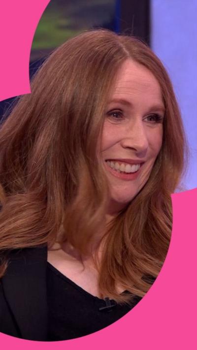 Catherine Tate has long red hair. She wears a black top and is smiling.