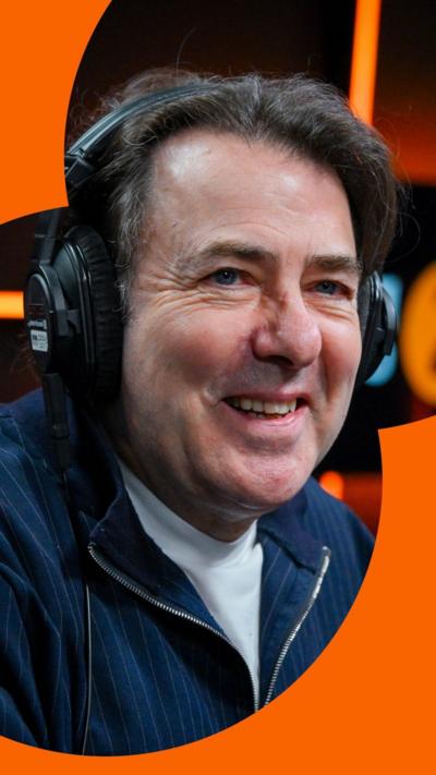 Broadcaster Jonathan Ross smiling with headphones on. He is wearing a blue jacket and white T-shirt.