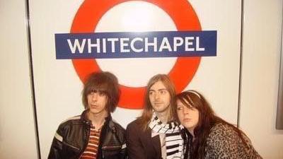 Three people pictured looking to the left at Whitechapel Tube station. The man on the left has long brown hair with a fringe. He wears a striped top and leather jacket. The man in the middle has shoulder length blonde hair and wears a piano style scarf and brown blazer. A girl rests her head on his shoulder and has brown fringed hair. She wears a leopard print top.