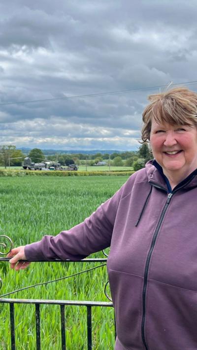 woman in a purple top stood by a field gate with a military helicopter in the distance