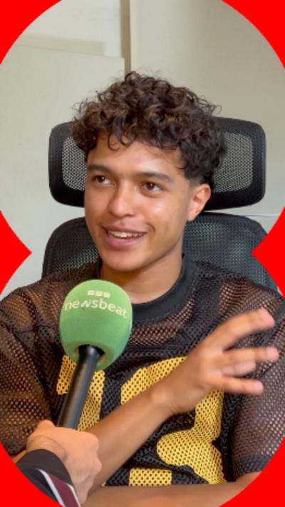 A young man with short curly hair. He's smiling and has a green microphone in front of him.