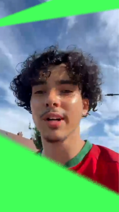 Young man with dark curly hair and red football shirt with blue sky and wispy clouds behind.