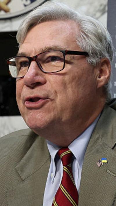 An old man with white hair, glasses and a tan suit speaks from a dais.