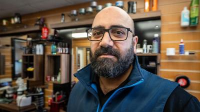 Mounzer Darsani, a barber originally from Syria, stands in his barbershop in the centre of Rothesay on the Isle of Bute. He has a black beard and black glasses and is wearing a dark blue waistcoat. In the background mirrors, scissors and hair products can be seen on shelves.