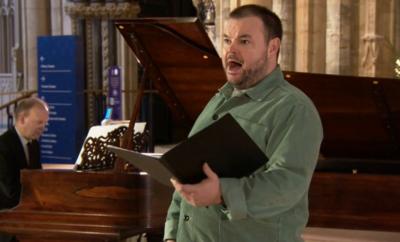 A man with short dark hair and dark-coloured stubble singing in a Church. He is wearing a long sleeve green shirt and is holding a folder open with his left hand. A man in a black suit is playing a piano behind him.