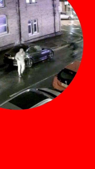 A man walking across the road in a grey tracksuit. There are cars on either side of the road, parked up.