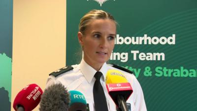 Gillian Kearney speaking at a police press conference. There is a green PSNI banner behind her and four microphones in front. She is in uniform and has blonde hair, which is tied back.