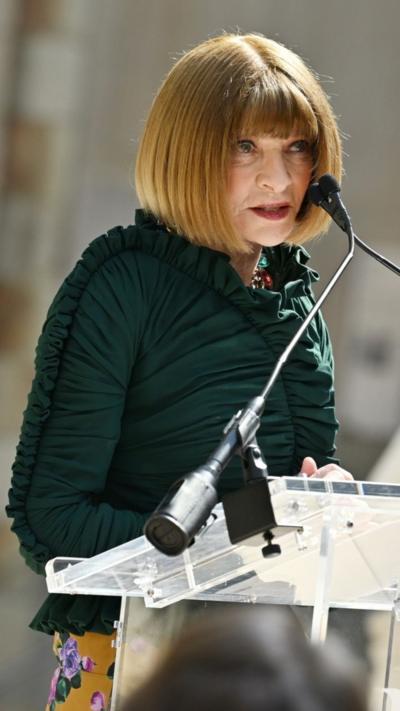 Anna Wintour, in a emerald green ruched top, speaks off camera towards her left into a microphone.
