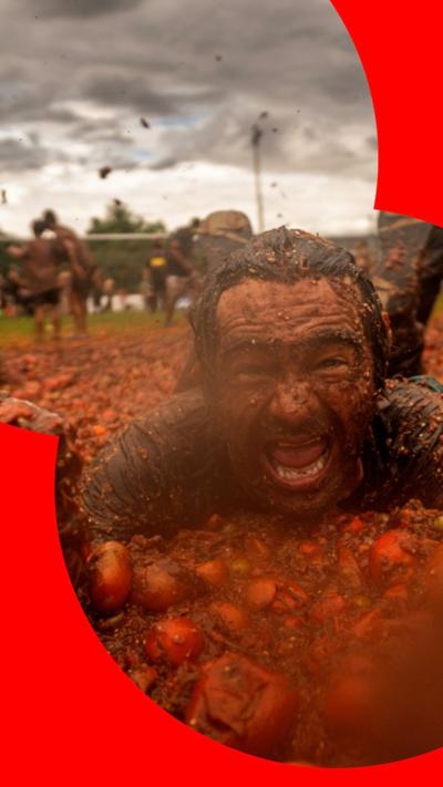 Man covered in tomato sauce lies in pile of tomatoes smiling at camera