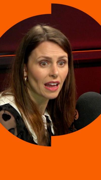 A woman, Ellie Taylor, has long brown hair and wears earrings. She speaks into a microphone