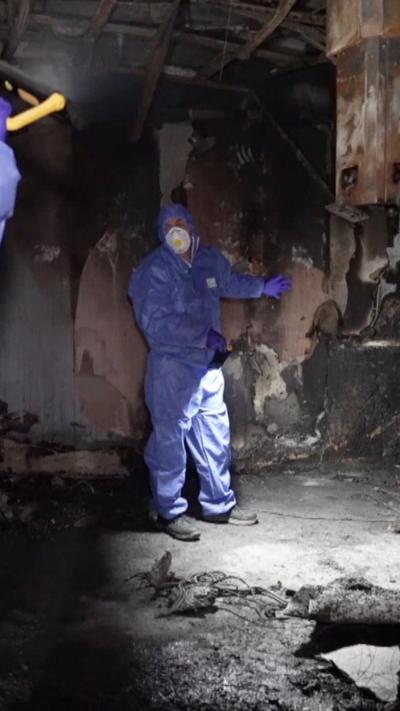 Man in blue PPE stands in fire-damaged room.