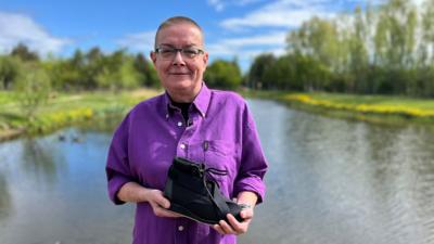 Jeanette Russell smiles and she stands in front of a body of water. It's a sunny day and there is lush green grass on either side with a row of trees and hedges on either side. She has cropped hair and is wearing glasses and a bright purple shirt. She is holding the black lace-up shoe.