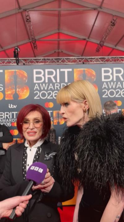 Two women being interviewed on the red carpet at the Brit Awards. The woman on the left has purple hair, glasses and a black suit with white shirt. The woman on the right has blonde hair and a feathered black top
