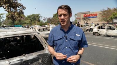 BBC correspondent in Mexico standing in front of a burnt car with a Costco store visible in the background
