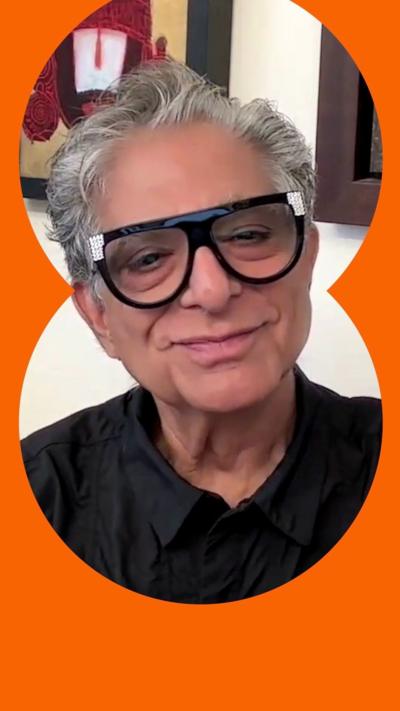 Deepak Chopra is wearing a black shirt and large black framed glasses.