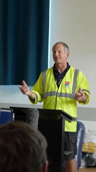 A man with short grey hair, wearing a yellow high-vis jacket, gesticulates as he faces a crowd in a hall.