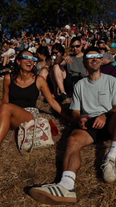 Two people smiling sat on a hill wearing eclipse glasses