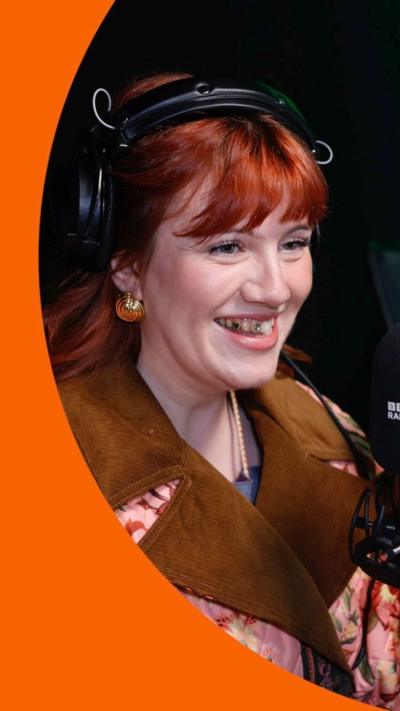 A woman, CMAT, in the BBC 6 Music studio. She has red hair, a pink floral coat and smiles, revealing some tooth gems