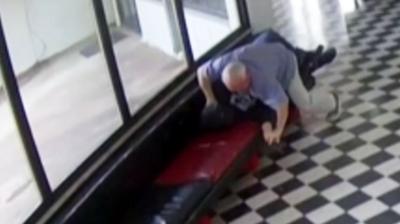 A still from CCTV footage showing a male in light coloured clothing lunging towards another male in dark clothing.