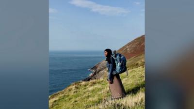 A woman is to the right of the photo and looking out to the sea. She is stood on a grassy hill. She is wearing a black hijab, blue jacket and brown skirt and has a blue backpack on.