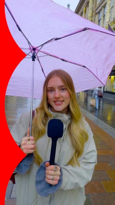 A woman stands in the street holding a microphone in one hand and a purple umbrella in the other.