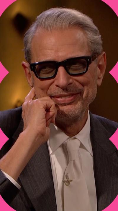 Jeff Goldblum smiles. He is wearing a white shirt and tie with a dark grey jacket, and black glasses with thick rims.