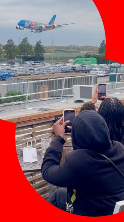 A colourful airplane descends towards an airport while onlookers take photos from a nearby observation area.