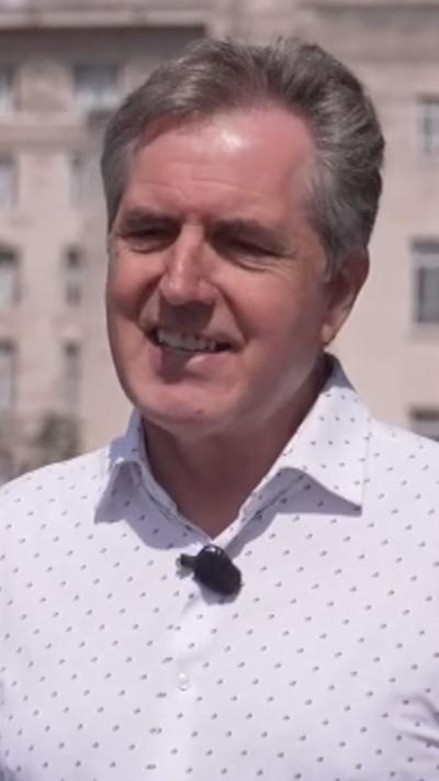 Steve Rotheram is in a white patterned shirt thats unbuttoned at the top and is smiling. He is stood outside facing the reporter.