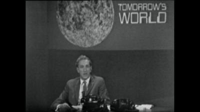 Tomorrow's World presenter sits at a desk with phones, below a sign with the Moon and 'Tomorrow's World' on it