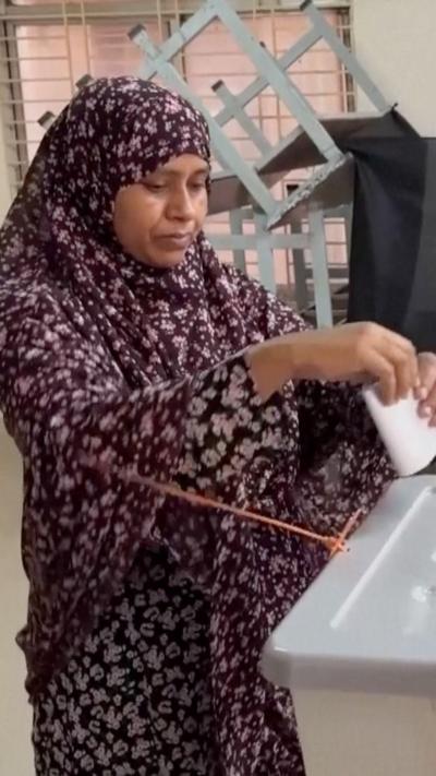A woman in a traditional purple dress inserting her paper vote into a container at a polling booth in Bangladesh