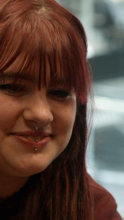 A woman with long brown hair and wearing a nose ring looks forward and smiles