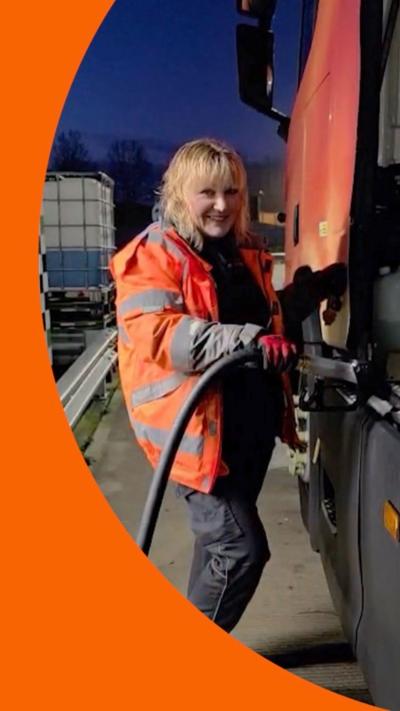 Woman with blonde hair and orange high vis jacket smiles while filling lorry with fuel.