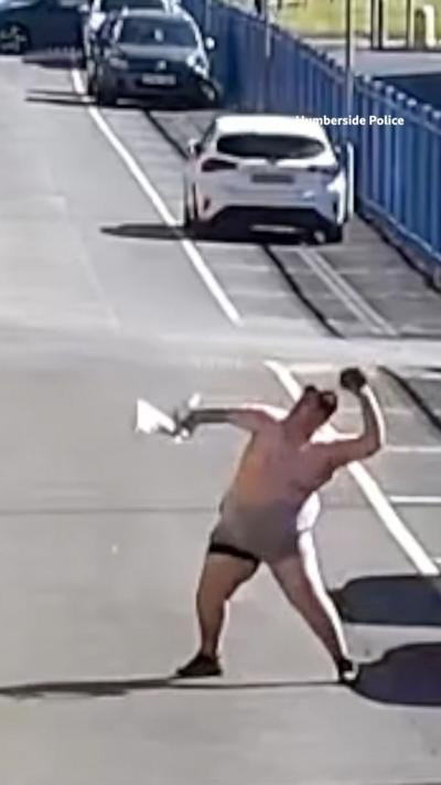 A bare-chested man wearing shorts stands in a street and leans back as he prepares to throw an object into the air.