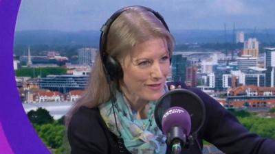 Donna Jones sits in the BBC Radio Solent studio with a backdrop of Southampton behind her. She has long blonde hair with headphones on and a navy blue knit jumper and silk floral scarf round her neck.