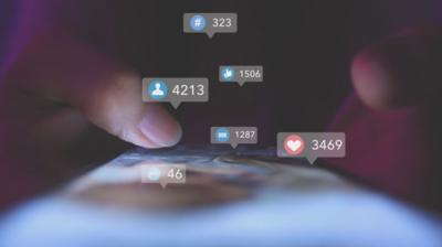 Two thumbs scrolling down a social media feed on a phone screen with likes, comments, shares and follow graphics surrounding it