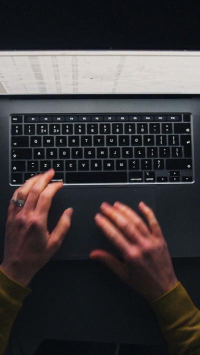Aerial view of hands typing on a laptop, with a cup of tea, a biscuit, and notepads filled with notes on a dark desk.