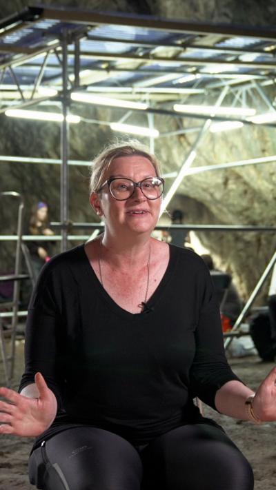A woman sat looking at the camera. She has glasses on and a black top and trousers. Behind her archaeological work is going on.