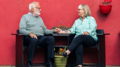 An older couple sits on a wooden bench against a bright red wall, holding hands across a green planter filled with flowers. The man has grey hair and a full beard, and wears a light green sweater and dark trousers, while the other woman wears a green‑striped shirt and black trousers. A red hanging pot with flowers is attached to the wall above them.