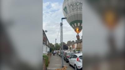 Hot air balloon in the sky seen approaching residential street. Houses on both sides and cars parked in front.