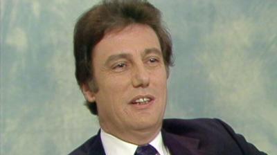 Paul Darrow, Black tie, white shirt, Black jacket, in a chatshow TV studio.